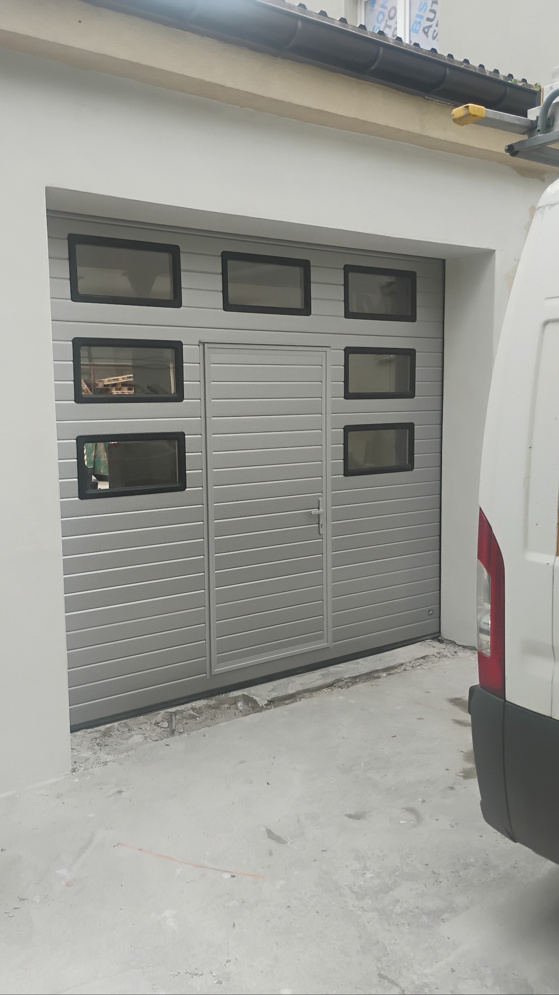Szara brama garażowa segmentowa z wbudowanymi drzwiami serwisowymi i przeszkleniami, widoczna z zewnątrz garażu. Częściowo widoczny biały samochód dostawczy.