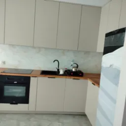 Beżowa zabudowa kuchenna z drewnianym blatem, czarnym zlewem i płytą indukcyjną, czarna bateria i czajnik na płycie, marmurowe wykończenie ściany nad blatem, częściowo zakryta folią lodówka po...