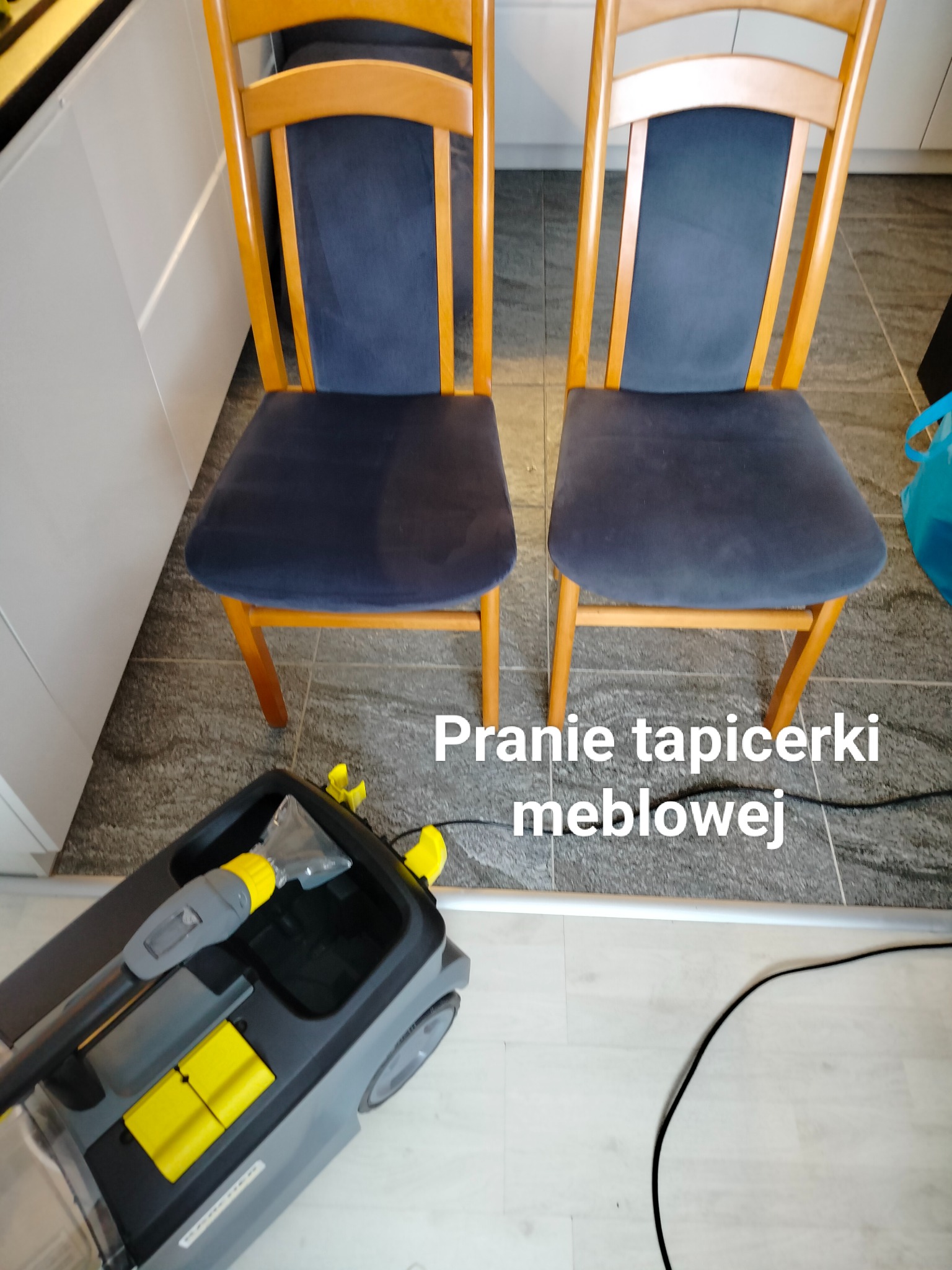 Dwa drewniane krzesła z granatową tapicerką siedzeń, obok szary odkurzacz piorący z żółtymi akcentami na szarej podłodze z płytek.