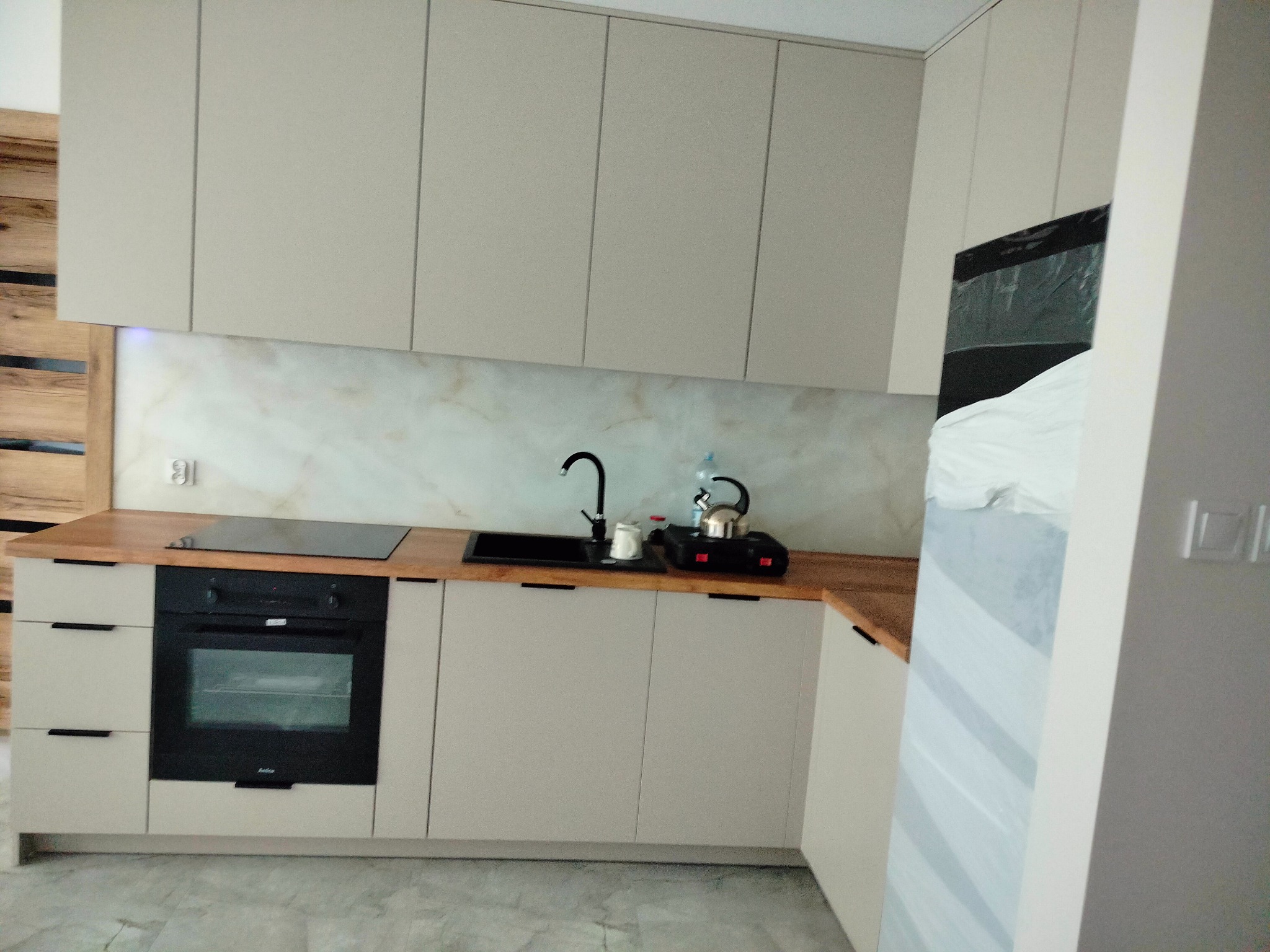 Beżowa zabudowa kuchenna z drewnianym blatem, czarnym zlewem i płytą indukcyjną, czarna bateria i czajnik na płycie, marmurowe wykończenie ściany nad blatem, częściowo zakryta folią lodówka po...