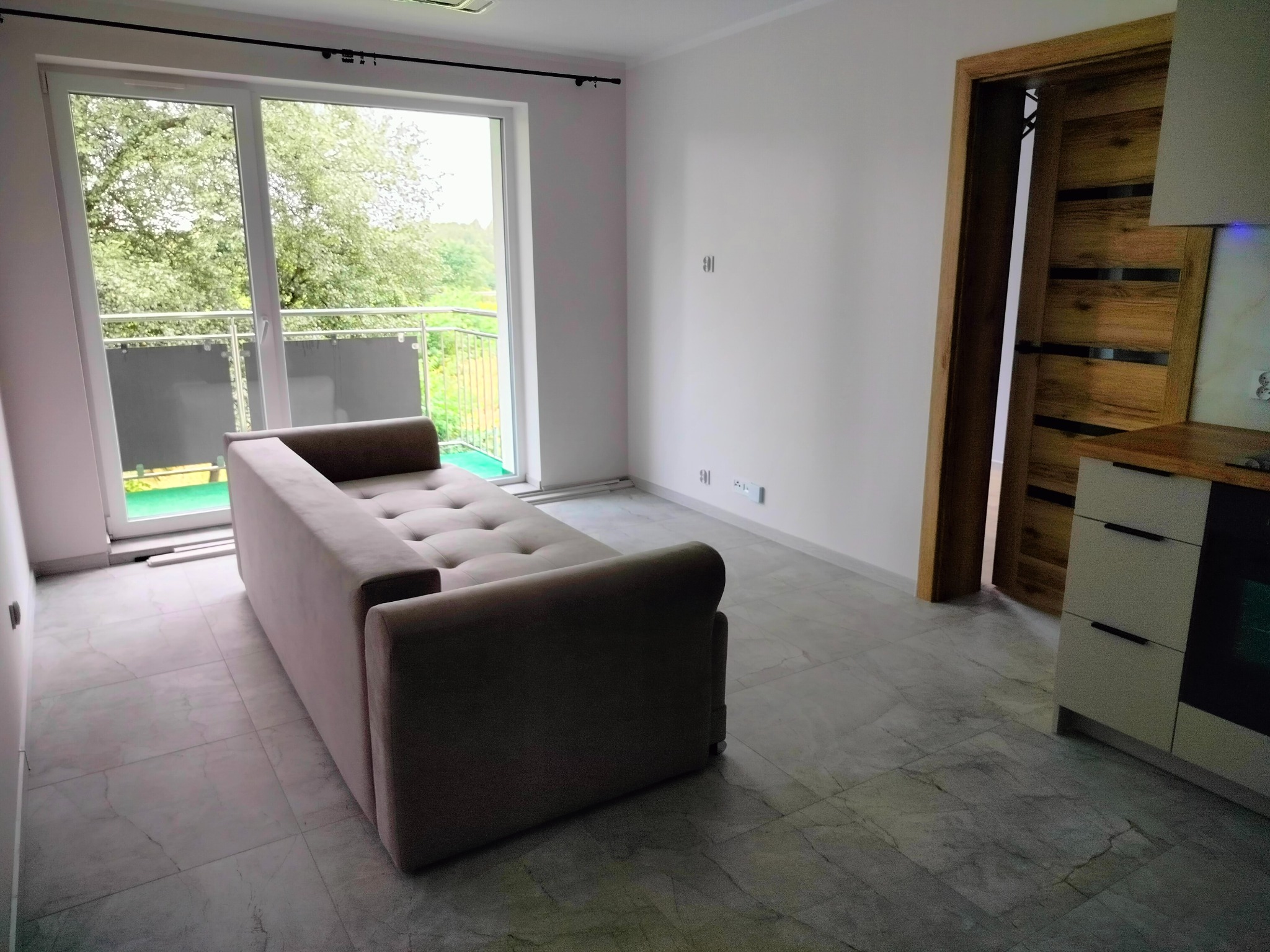 Pusta, jasna przestrzeń z szarą sofą na marmurowej podłodze, balkon z widokiem na zieleń i drewniane drzwi w tle.