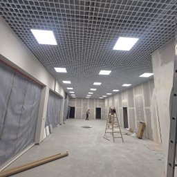 ELEKTRYKAS Tadeusz Suszczewicz - Sufit rastrowy z zamontowanym oświetleniem w przestrzeni komercyjnej w trakcie prac wykończeniowych. Widoczna drabina i osoba w oddali.
