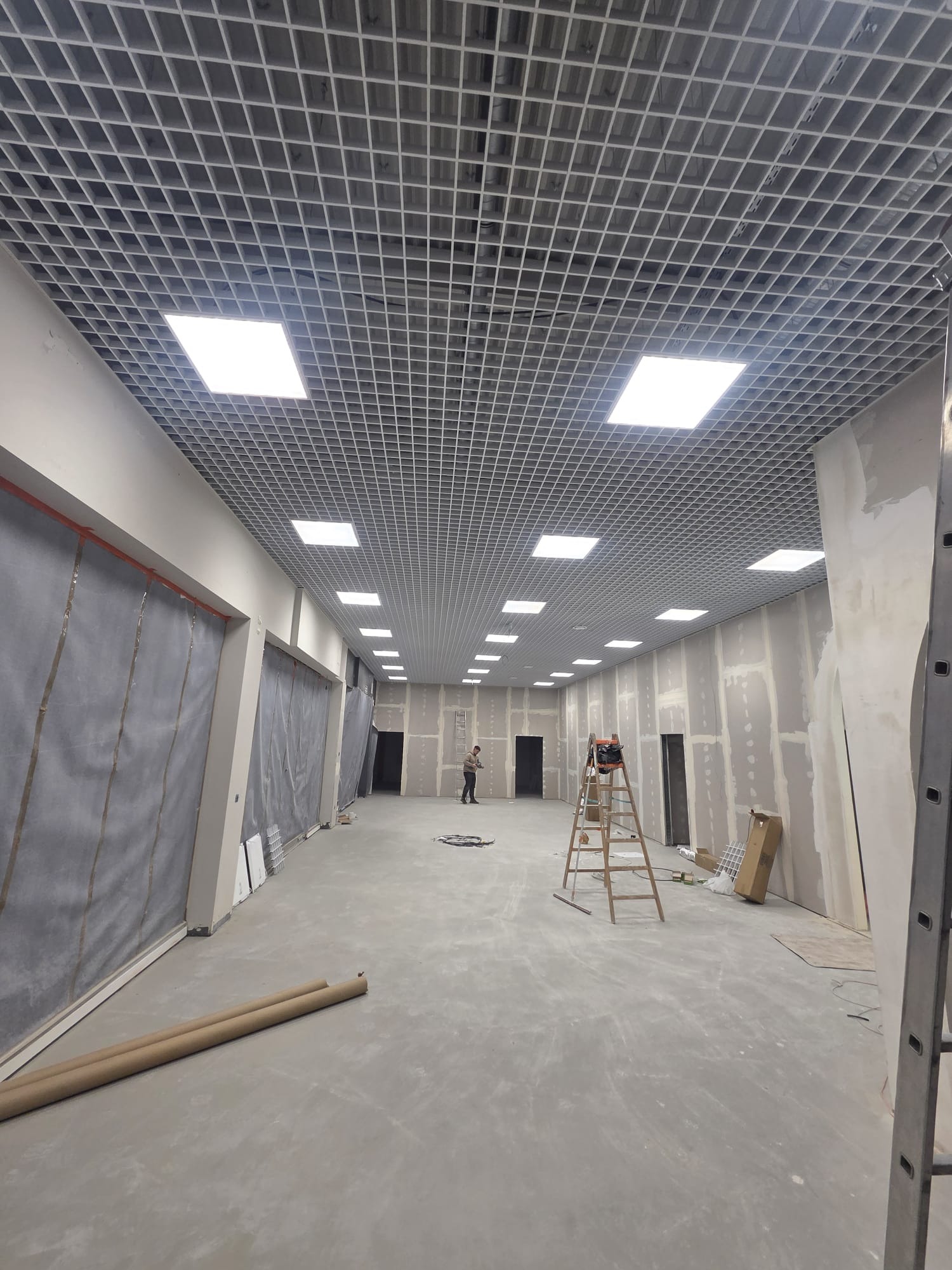 Sufit rastrowy z zamontowanym oświetleniem w przestrzeni komercyjnej w trakcie prac wykończeniowych. Widoczna drabina i osoba w oddali.
