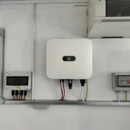 Biały inwerter fotowoltaiczny firmy Huawei zamontowany na ścianie z dwoma skrzynkami elektrycznymi, połączony przewodami w kolorach czerwonym, białym i zielonym, umieszczony na metalowej szynie...