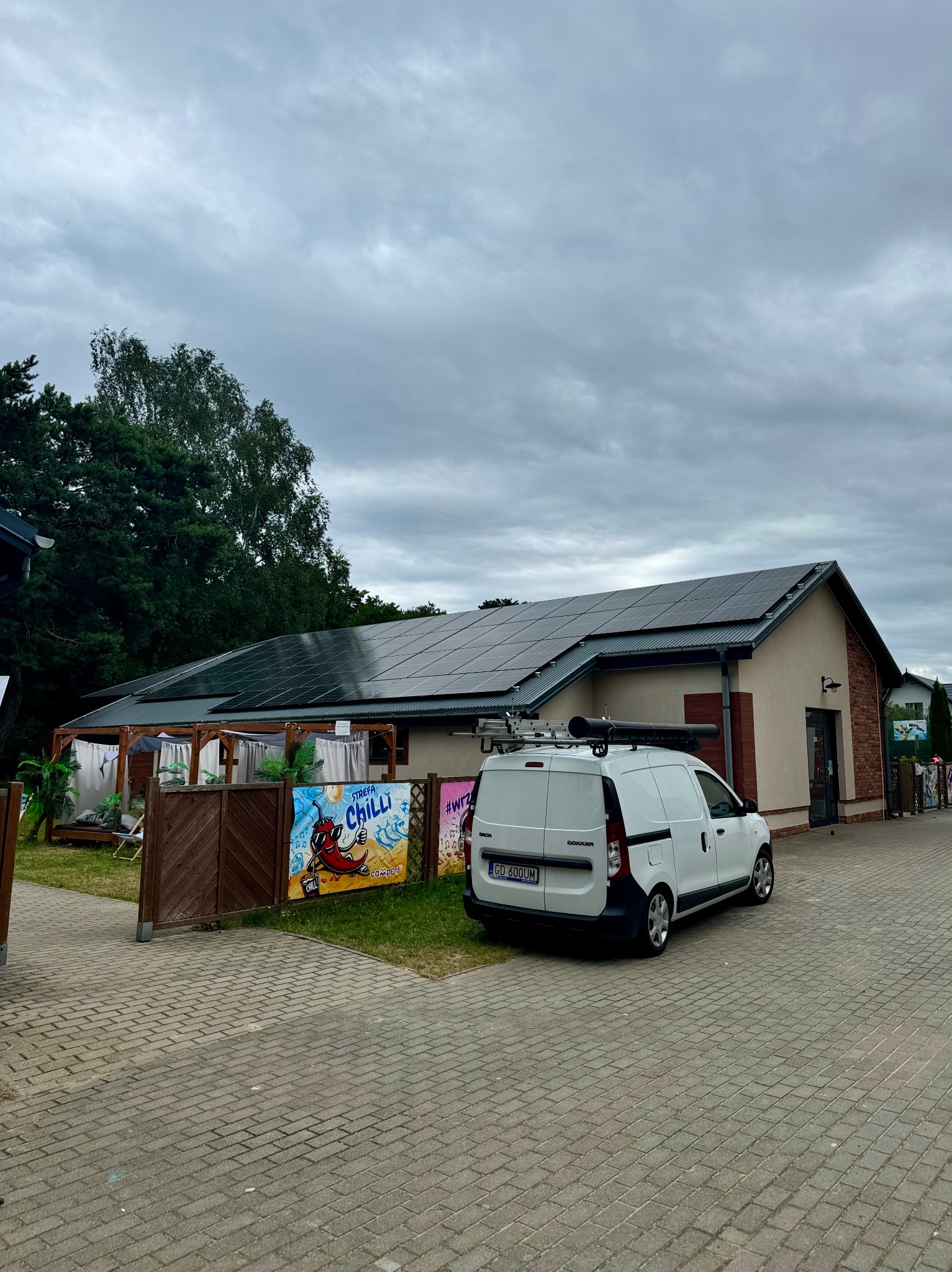 Budynek z panelami słonecznymi na dachu, obok zaparkowany biały samochód dostawczy z bagażnikiem dachowym, widoczne reklamy na ogrodzeniu.