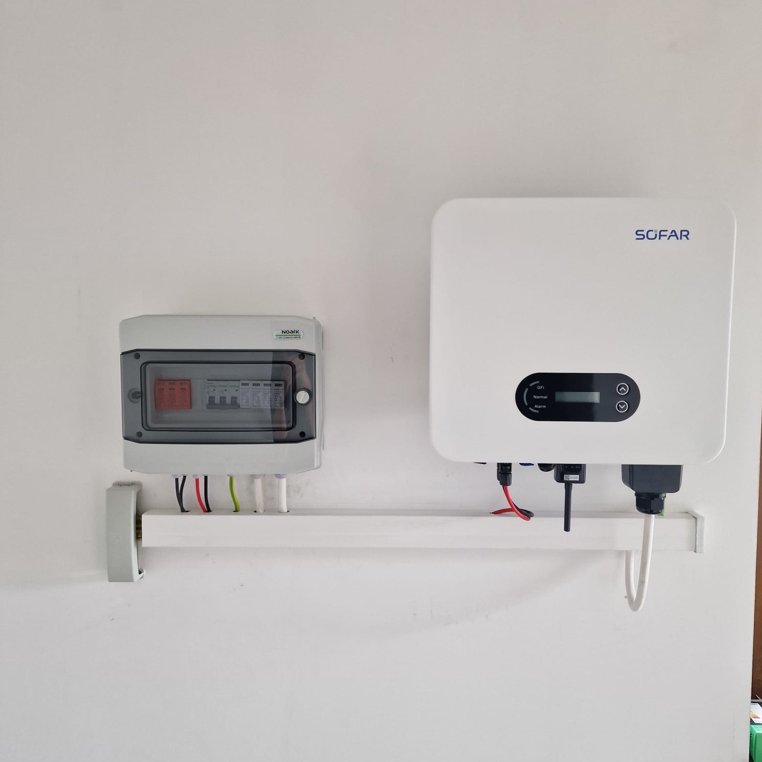 Biały falownik Sofar z podłączonymi kablami, obok szara skrzynka z bezpiecznikami na białej ścianie, zamontowane na białym wsporniku.