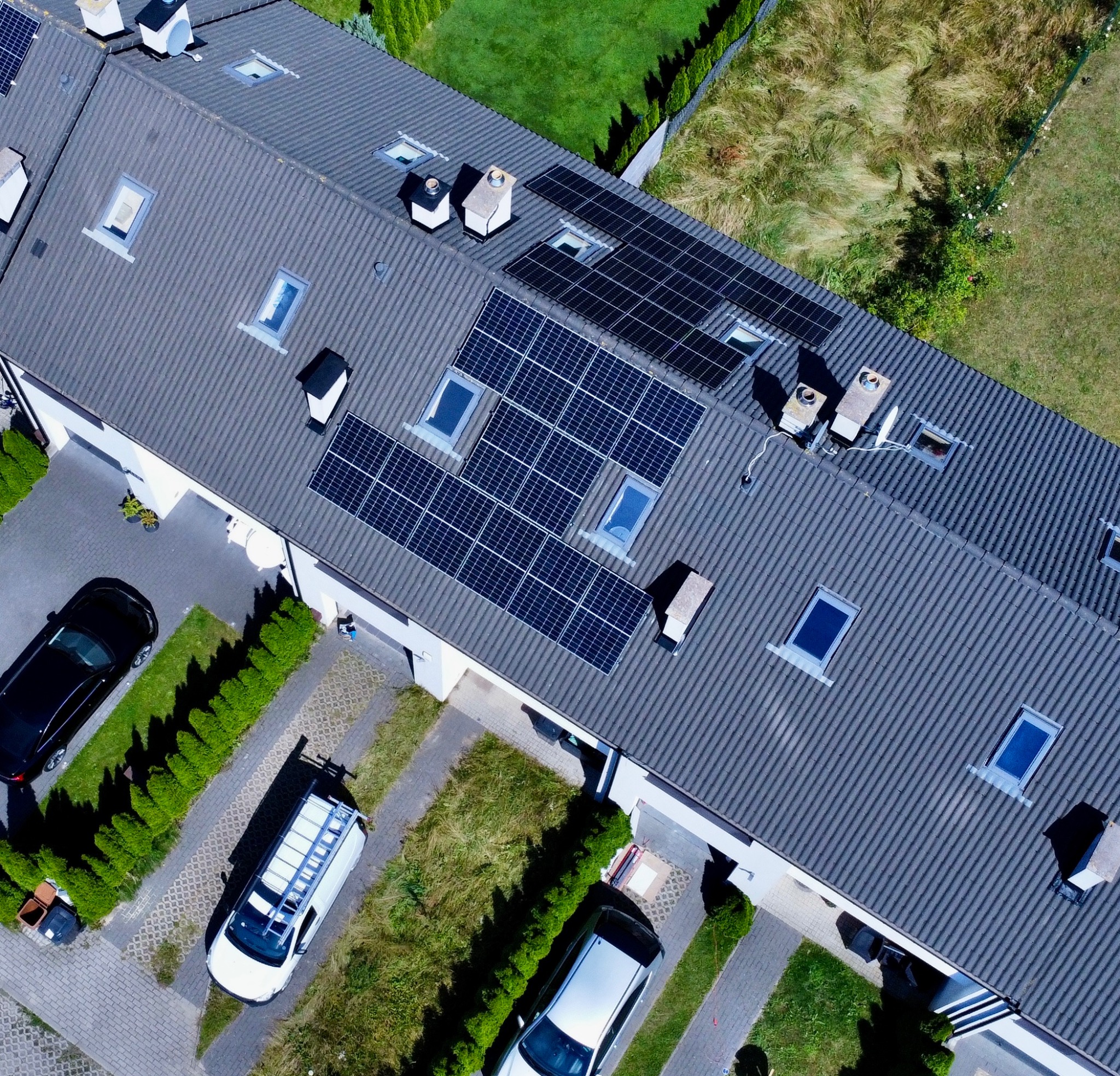Widok z góry na szereg domów z zainstalowanymi panelami fotowoltaicznymi na dachach, zaparkowane samochody i bus z drabiną.