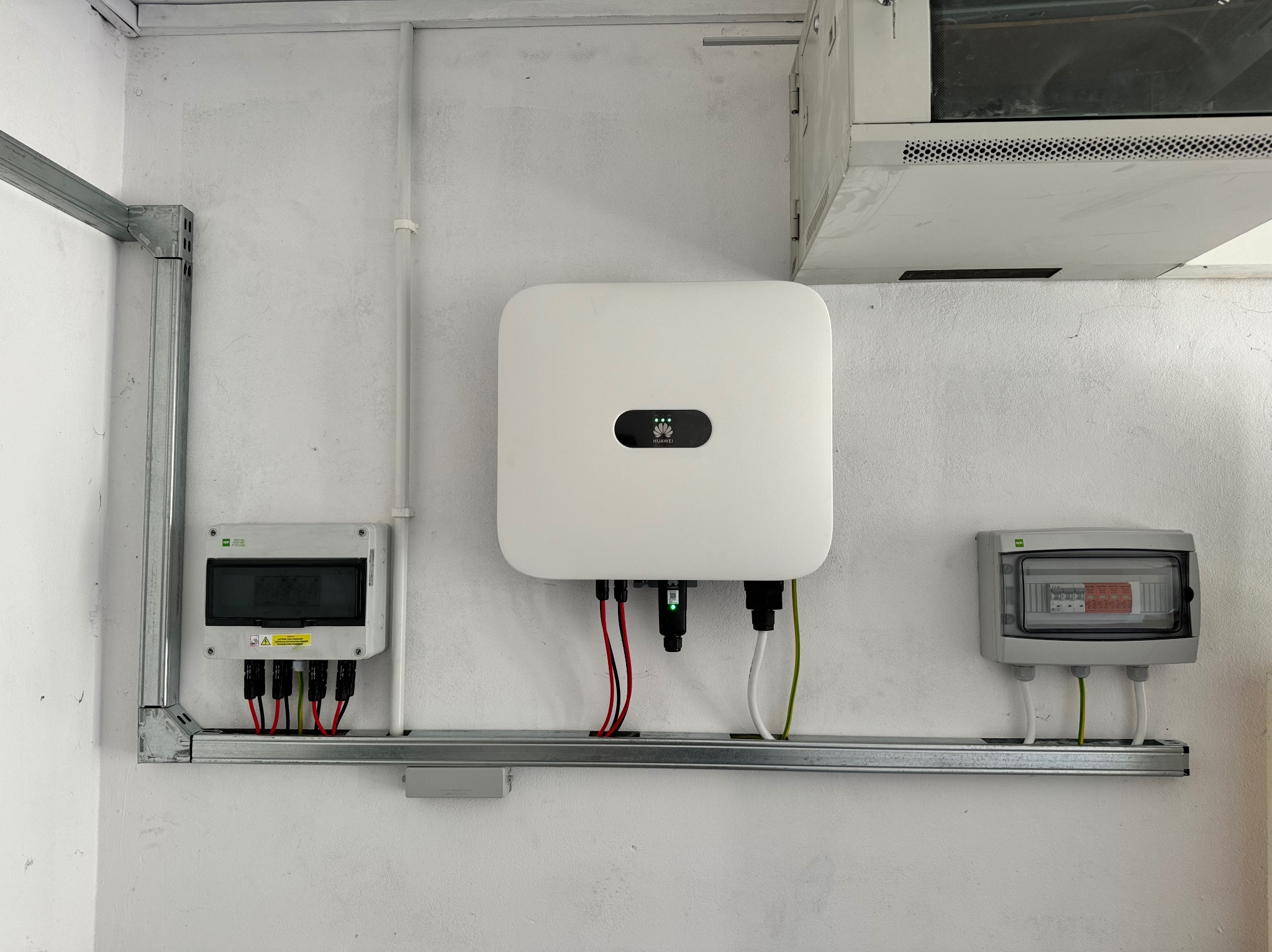 Biały inwerter fotowoltaiczny firmy Huawei zamontowany na ścianie z dwoma skrzynkami elektrycznymi, połączony przewodami w kolorach czerwonym, białym i zielonym, umieszczony na metalowej szynie...