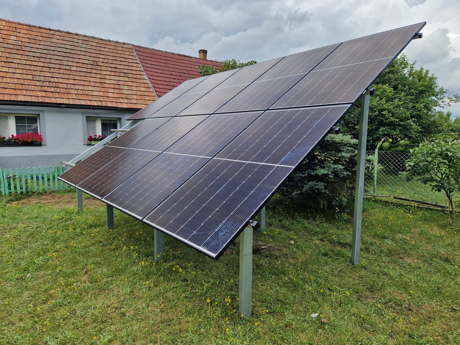 Panel słoneczny na metalowych słupkach w ogrodzie, z domem z czerwonym dachem i zielonym płotem w tle, pochmurne niebo.