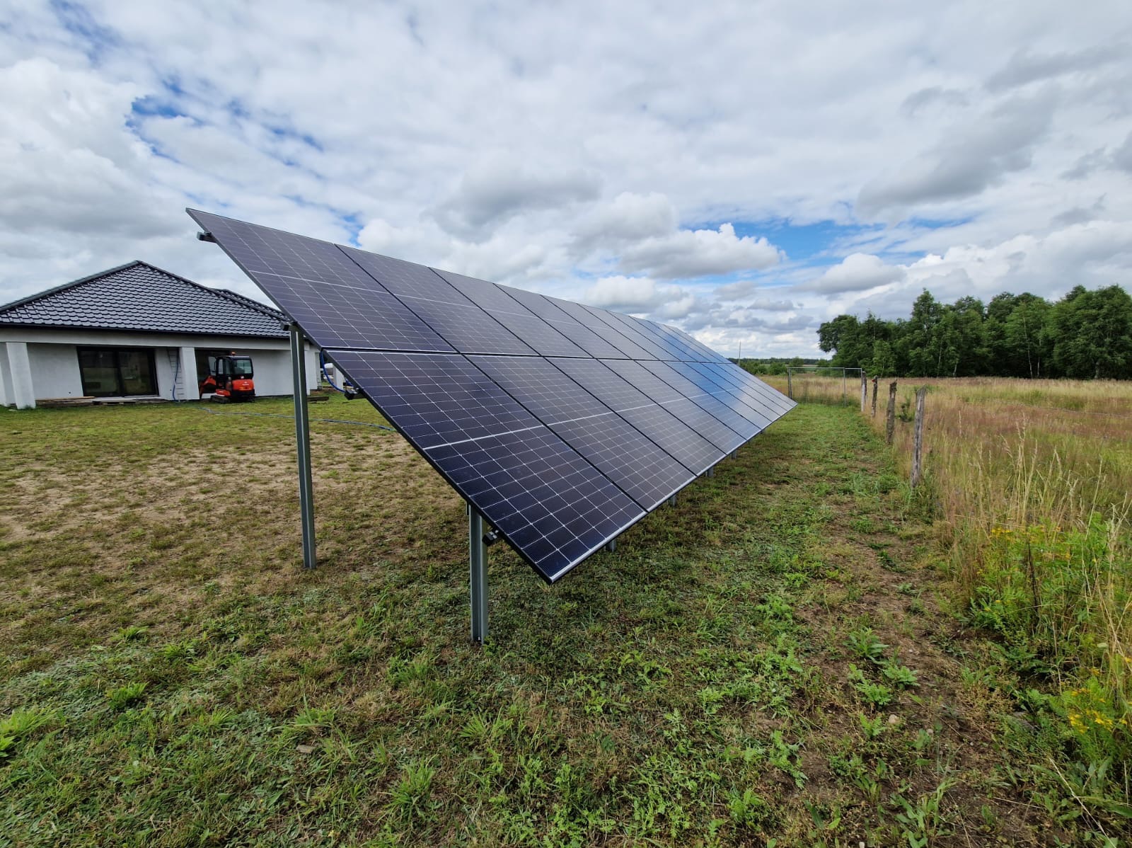 Panel słoneczny zamontowany na metalowych podporach na trawiastym terenie, z domem i minikoparką w tle, widoczne pochmurne niebo.