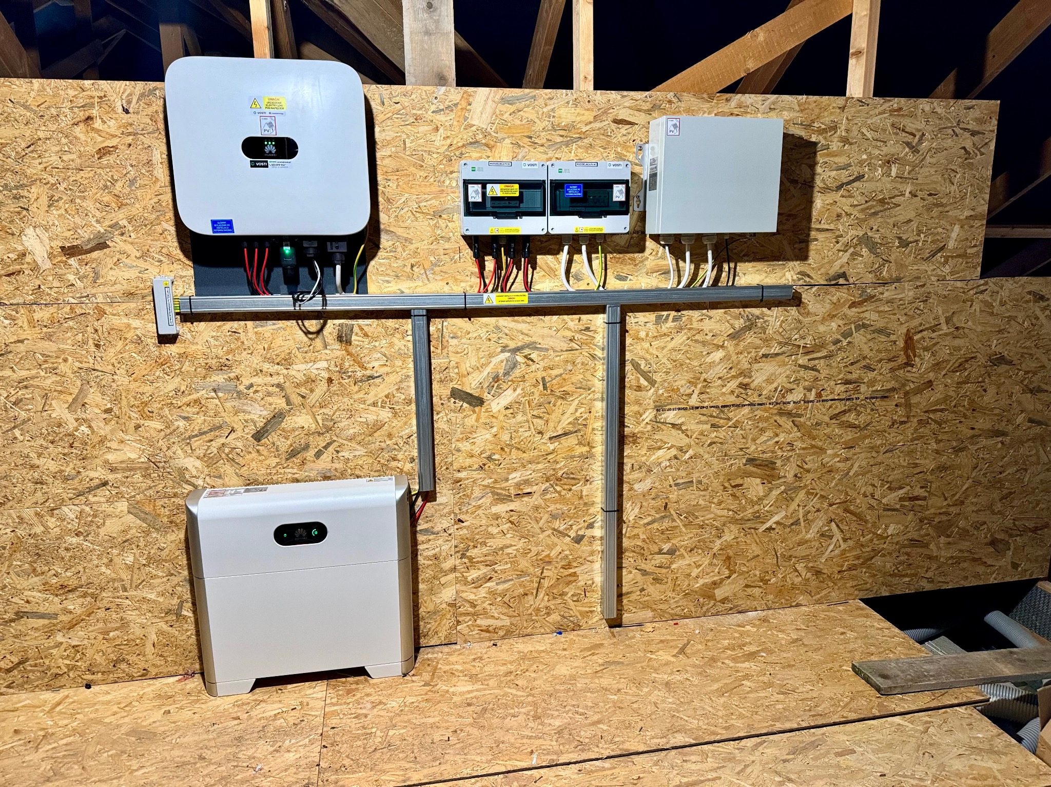 Zamontowany inwerter fotowoltaiczny i magazyn energii na ścianie z płyty OSB w poddaszu, widoczne zabezpieczenia elektryczne i okablowanie.