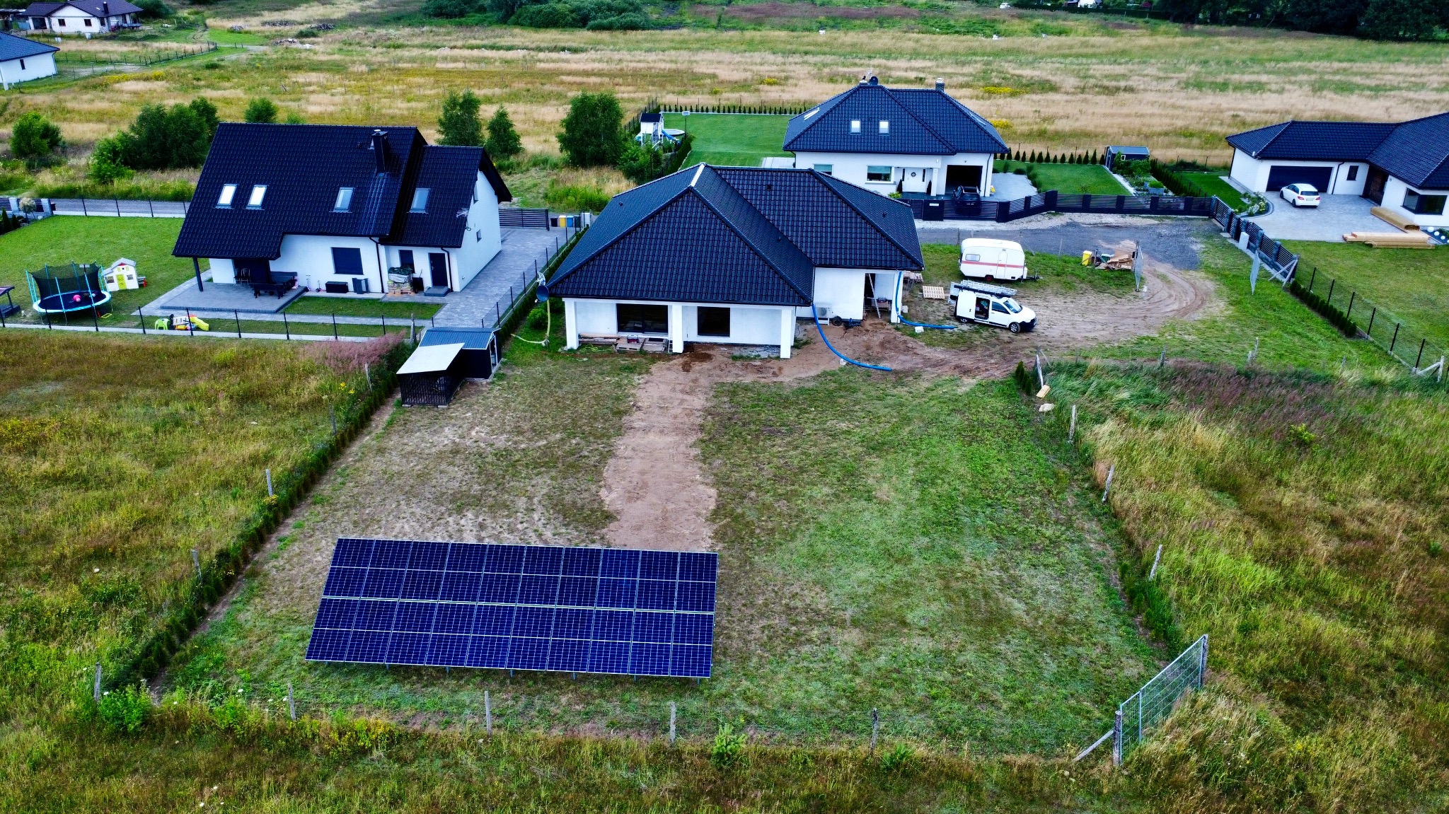 Widok z góry na panele słoneczne w ogrodzie przed domem jednorodzinnym, w tle inne domy i zieleń.