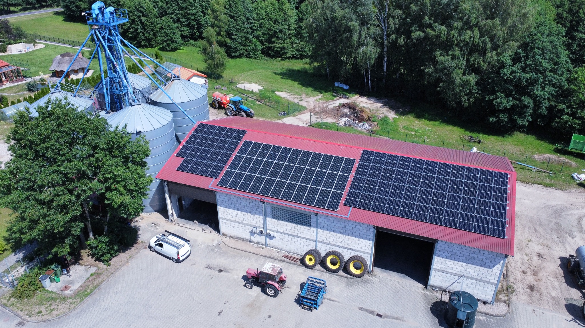 Widok z góry na budynek gospodarczy z czerwonym dachem pokrytym panelami fotowoltaicznymi, obok silosy zbożowe i maszyny rolnicze.