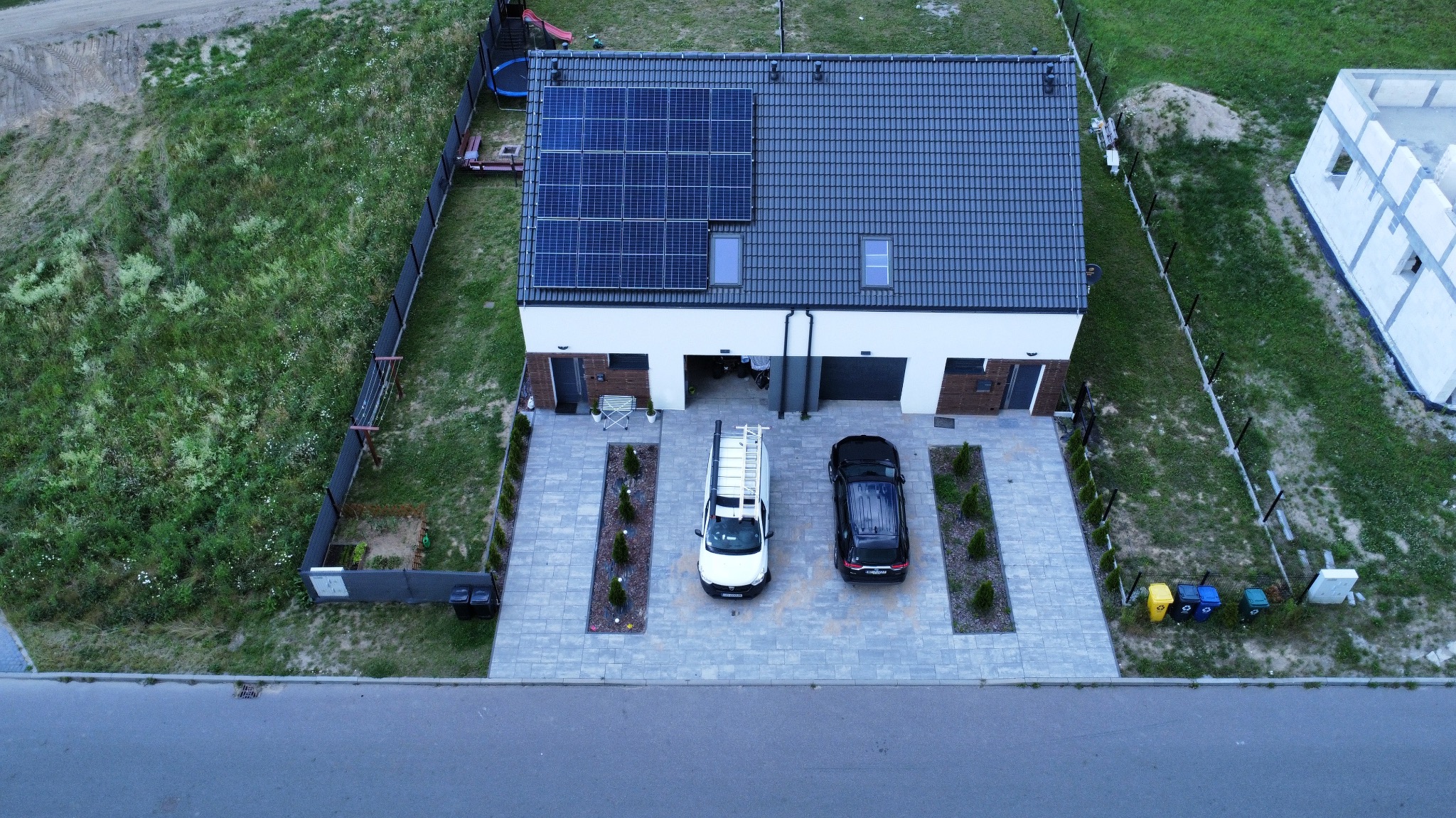 Widok z drona na nowoczesny dom z panelami słonecznymi na dachu, zaparkowany biały bus z drabiną i czarny samochód na brukowanym podjeździe, w tle ogród i budowa.
