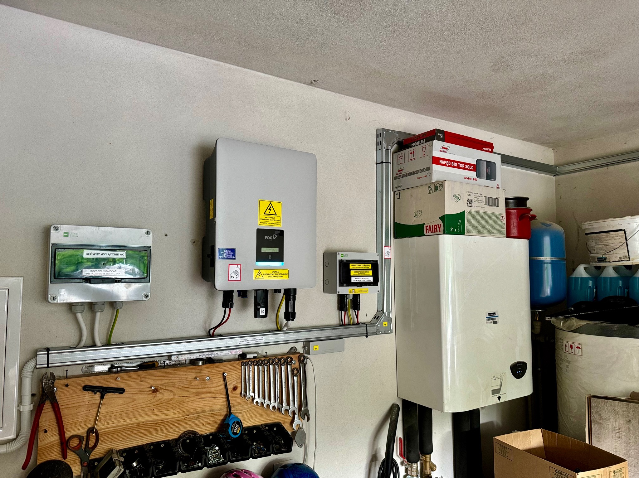 Szary inwerter fotowoltaiczny FoxESS z żółtą naklejką ostrzegawczą, zamontowany na ścianie nad drewnianą listwą z narzędziami, obok rozdzielnicy PV AC, z widocznymi kablami i przewodami elektrycznymi.