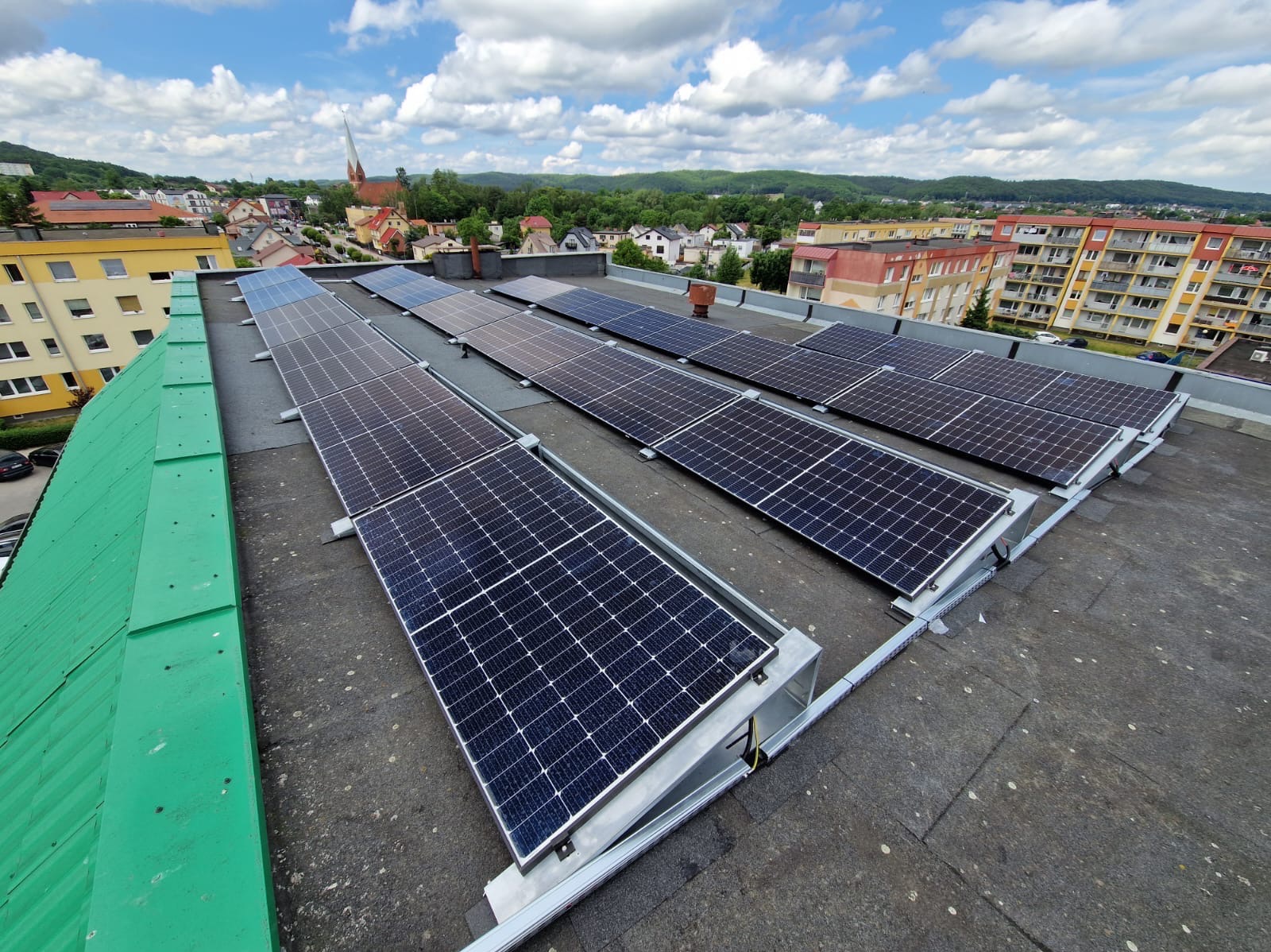 Rząd paneli słonecznych na płaskim dachu budynku z widokiem na miasto i kościół w tle, błękitne niebo z chmurami.