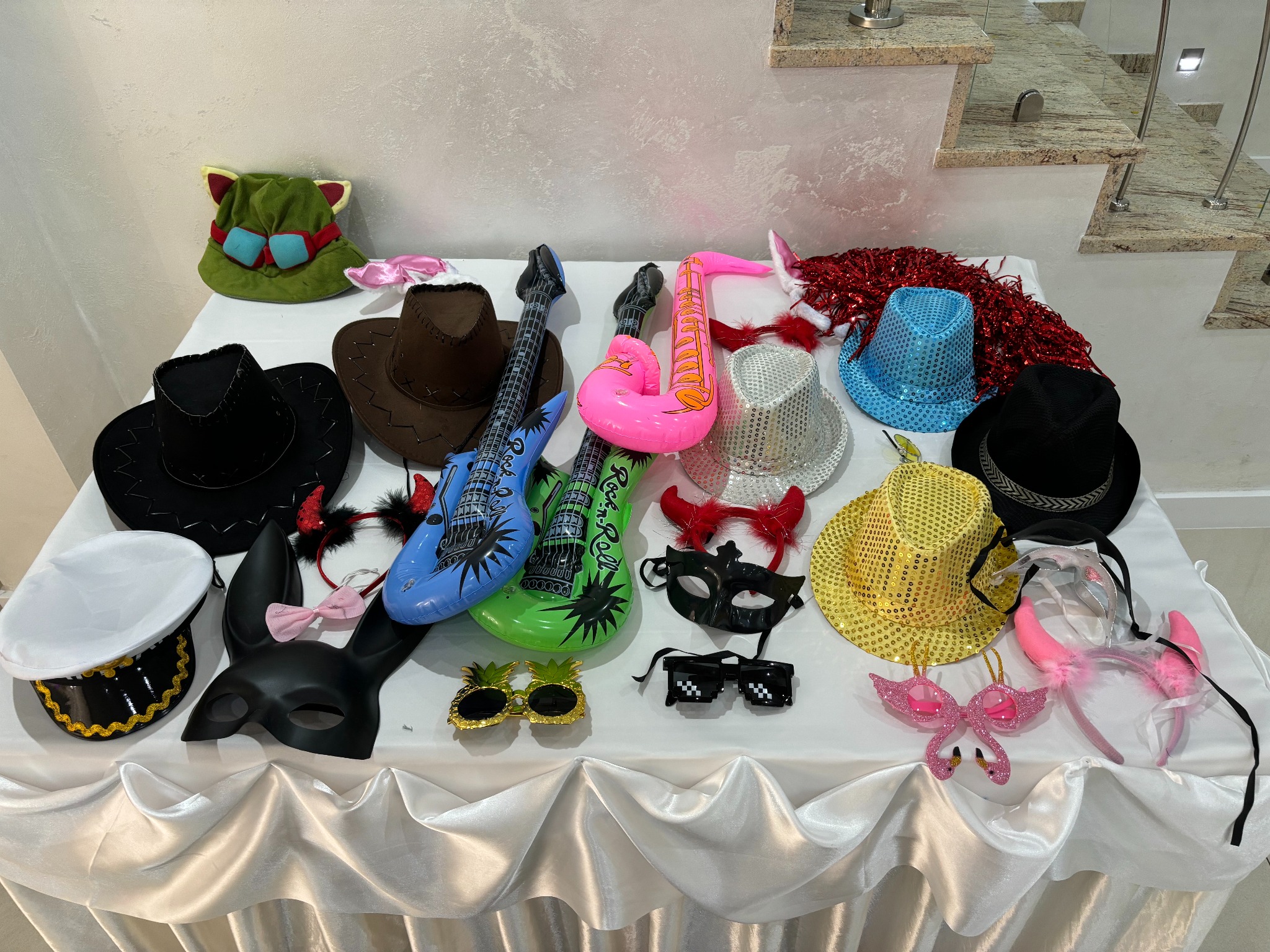 Różnorodny zestaw rekwizytów do zdjęć: kapelusze, maski, okulary, dmuchane instrumenty, leżące na białym stole z dekoracyjnym obrusem i schodami w tle.