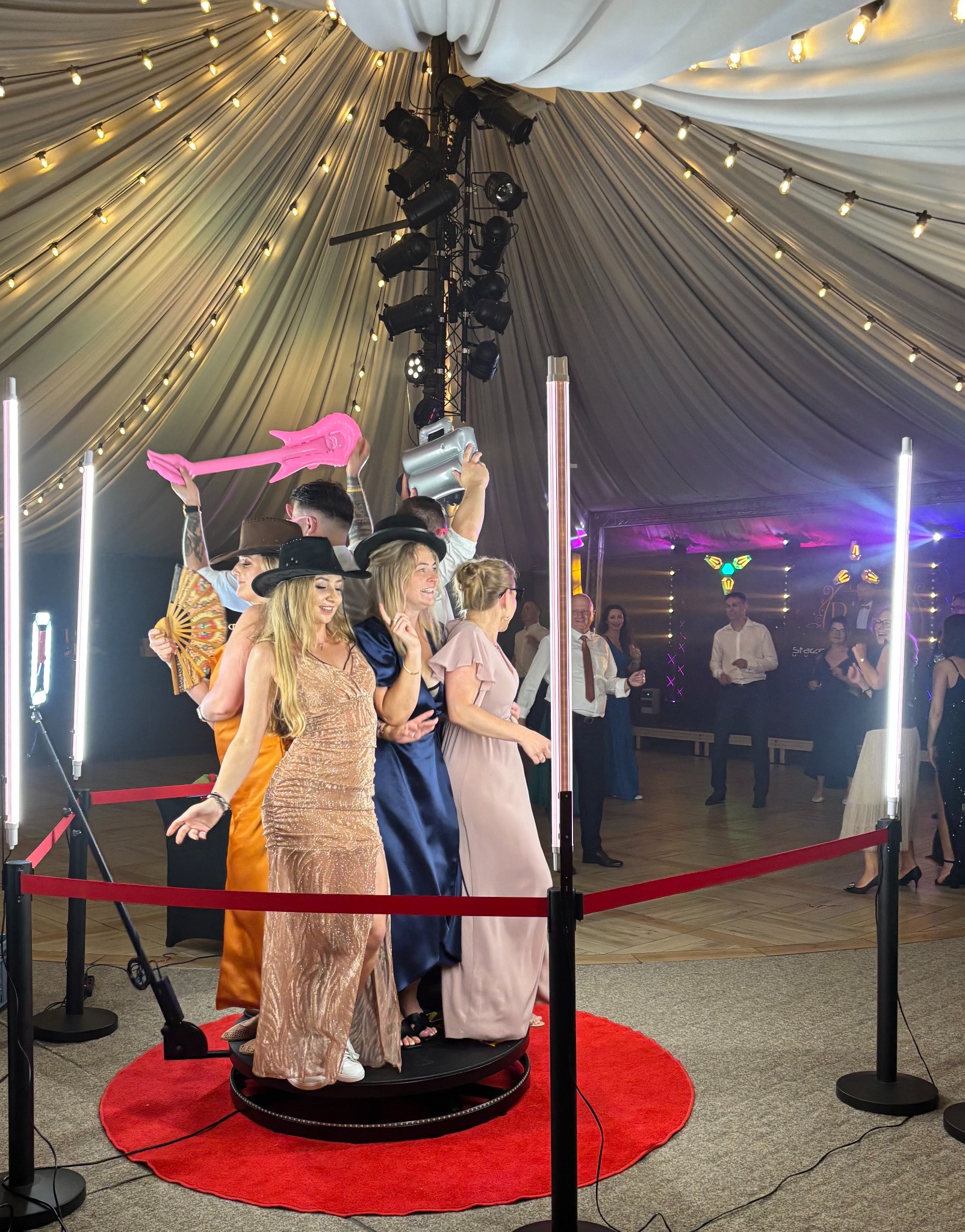 Grupa osób w eleganckich strojach pozuje z rekwizytami (dmuchana gitara, wiadro) w obrotowej fotobudce 360 stopni na tle dekoracji świetlnych.