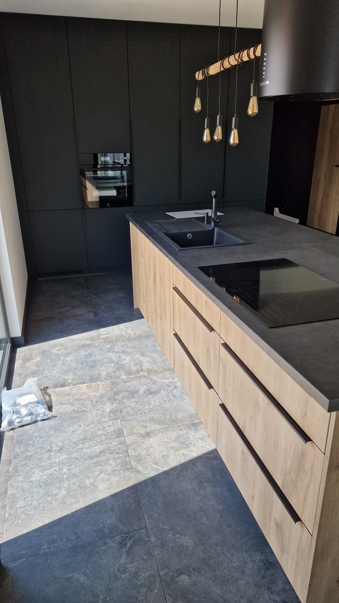 Nowoczesna kuchnia: czarne szafki na tle jasnego blatu wyspy z wbudowaną płytą indukcyjną i zlewozmywakiem, oświetlona wiszącymi lampami edisonkami, z widocznymi elementami montażowymi na podłodze.
