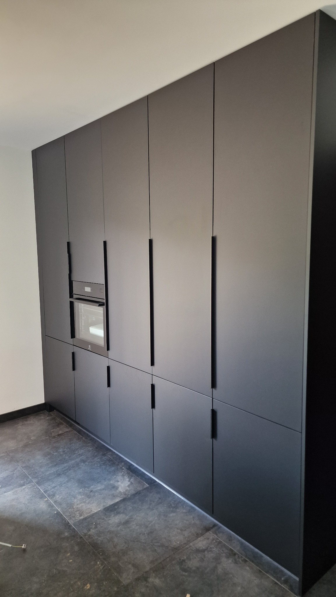 Ciemnoszara zabudowa kuchenna od podłogi do sufitu z piekarnikiem wbudowanym w szafkę i podłużnymi uchwytami.