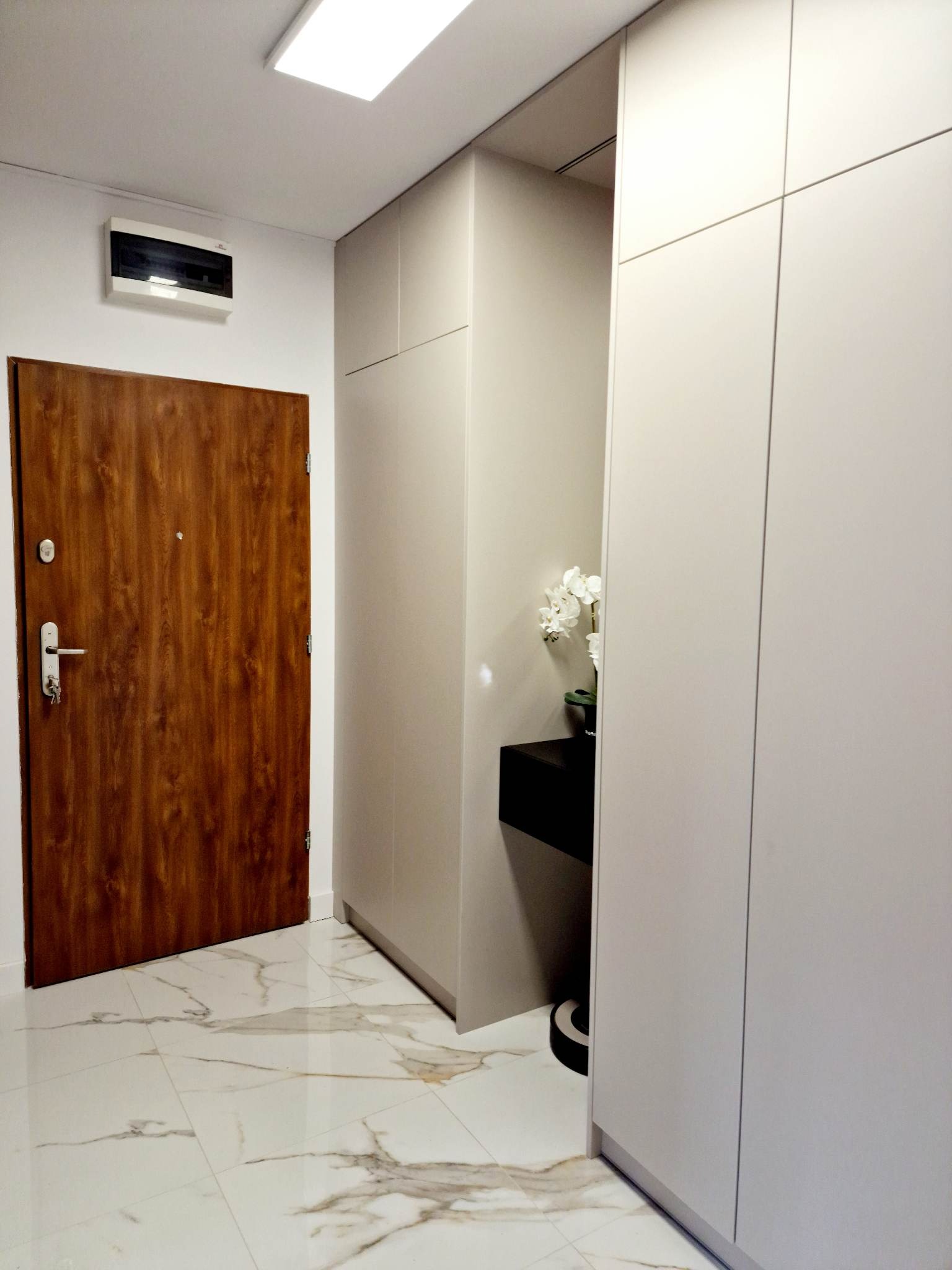 Korytarz z zabudowaną szafą w kolorze jasnego beżu, drewniane drzwi wejściowe, podłoga z białego marmuru z szarymi żyłami, czarna konsola z białymi kwiatami.
