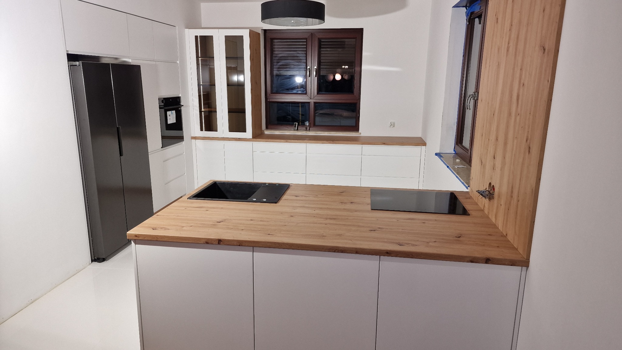 Nowoczesna kuchnia z białymi frontami, drewnianym blatem, czarnym zlewem i płytą indukcyjną, oraz dużą lodówką. Widoczne okno z brązową ramą i żaluzjami.