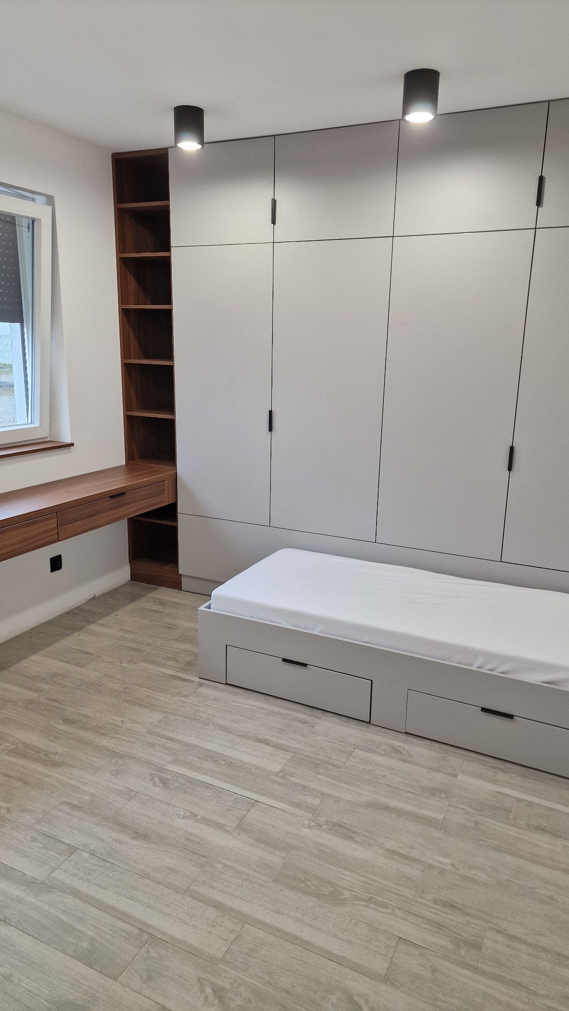 Szafa wnękowa z szarymi frontami, zintegrowana z łóżkiem z szufladami i biurkiem z półkami z drewna orzechowego, oświetlenie punktowe, jasna podłoga imitująca drewno.