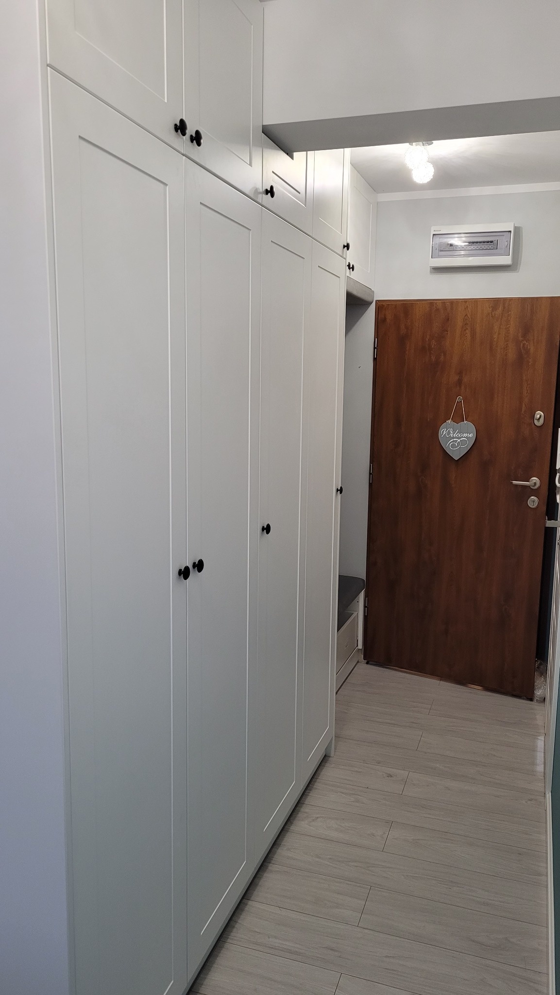 Biała szafa wnękowa z czarnymi uchwytami, widoczna w przedpokoju z drewnianymi drzwiami i szarą podłogą imitującą drewno.