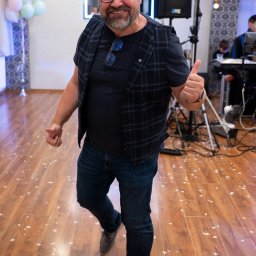 REBEL RECORDS FILM&FOTO RAFAŁ HEBEL - Uśmiechnięty kamerzysta w trakcie pracy na sali balowej, z uniesionym kciukiem, w tle balony, sprzęt nagłośnieniowy i zespół muzyczny.