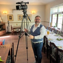 REBEL RECORDS FILM&FOTO RAFAŁ HEBEL - Mężczyzna w okularach i kamizelce stoi za kamerą wideo na statywie w sali z nakrytym stołem, gotowym do filmowania uroczystości.