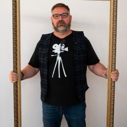 REBEL RECORDS FILM&FOTO RAFAŁ HEBEL - Mężczyzna z brodą i okularami, ubrany w koszulkę z rysunkiem kamery, trzymający ozdobną, złotą ramę.