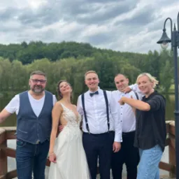 Panna młoda w koronkowej sukni i pan młody z muchą, pozujący z gośćmi na drewnianym pomoście z widokiem na jezioro i las, pochmurne niebo.