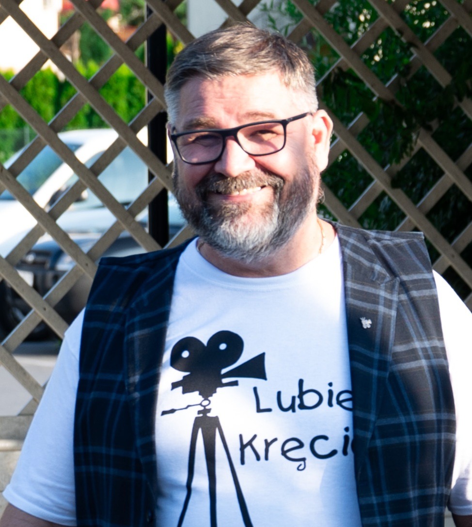 Uśmiechnięty mężczyzna w okularach i kamizelce, z koszulką z grafiką kamery i napisem 'Lubię Kręcić', na tle drewnianej kratownicy.