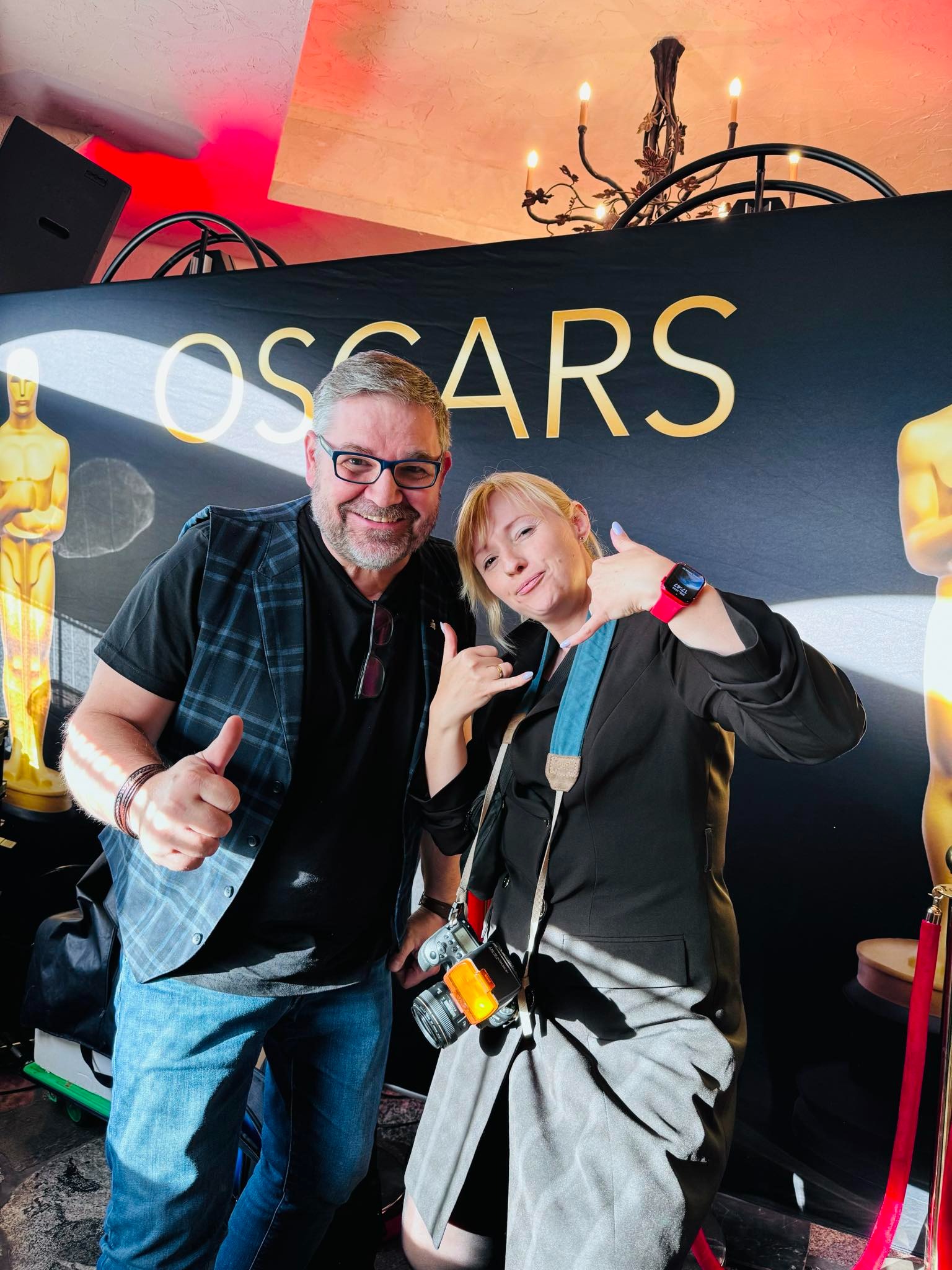 Dwie osoby pozują na tle plakatu z napisem OSCARS, kobieta z aparatem na szyi pokazuje gest 'hang loose', mężczyzna z brodą unosi kciuki w górę.