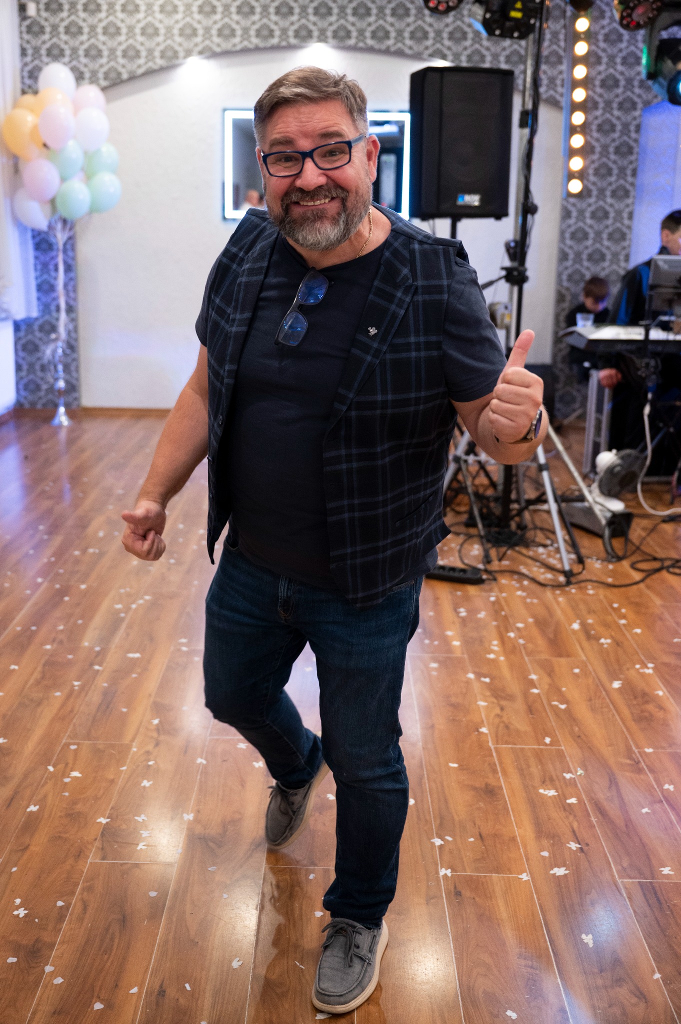 Uśmiechnięty kamerzysta w trakcie pracy na sali balowej, z uniesionym kciukiem, w tle balony, sprzęt nagłośnieniowy i zespół muzyczny.