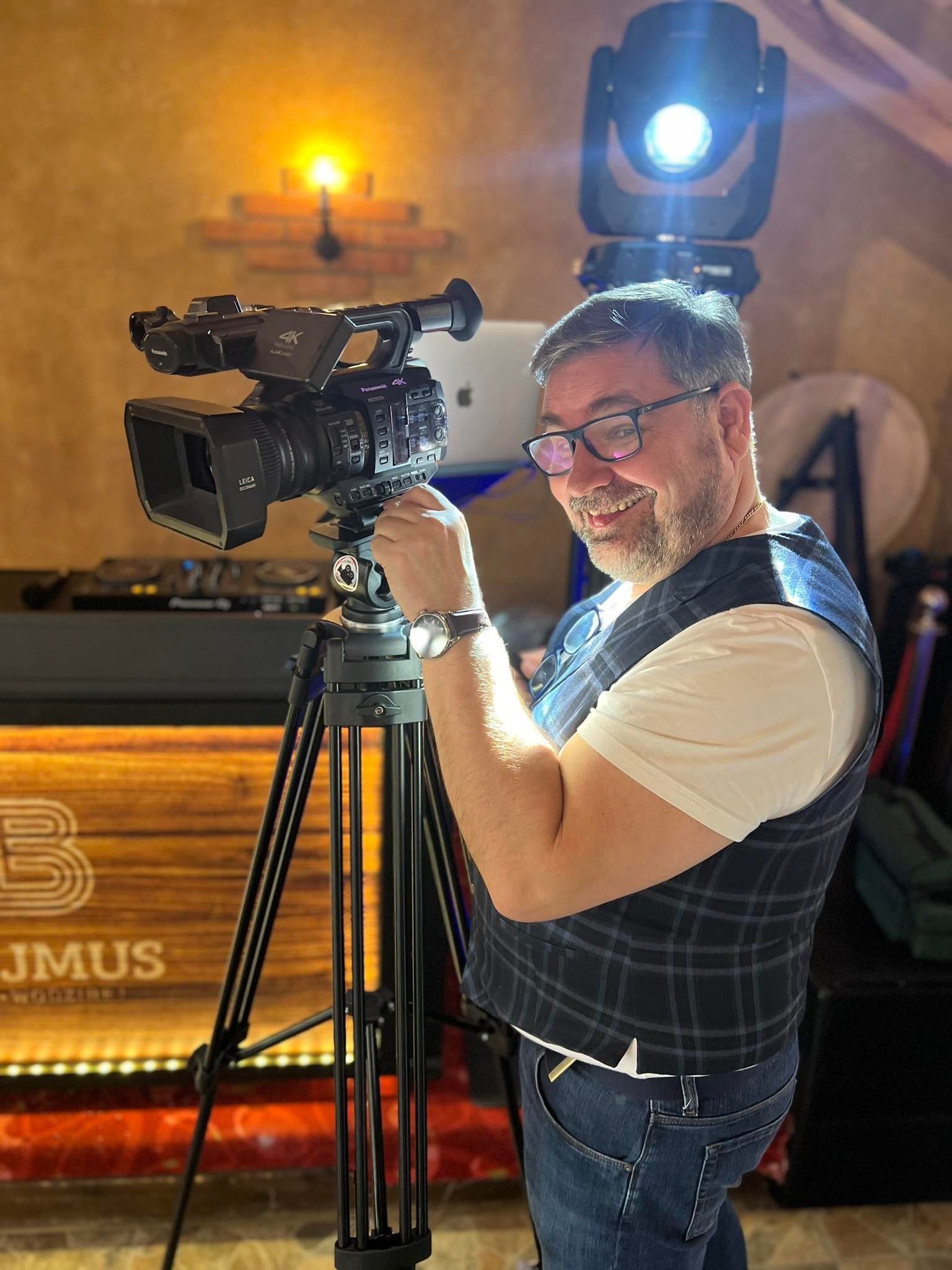 Uśmiechnięty mężczyzna w okularach i kamizelce w kratę, stojący za kamerą Panasonic 4K na statywie, z oświetleniem scenicznym w tle i konsolą DJ-ską.