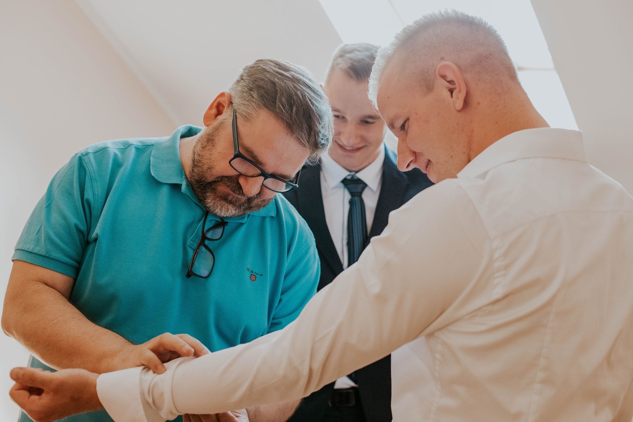 Przygotowania do ślubu: ojciec poprawia mankiet koszuli młodego pana, świadek uśmiecha się w tle.