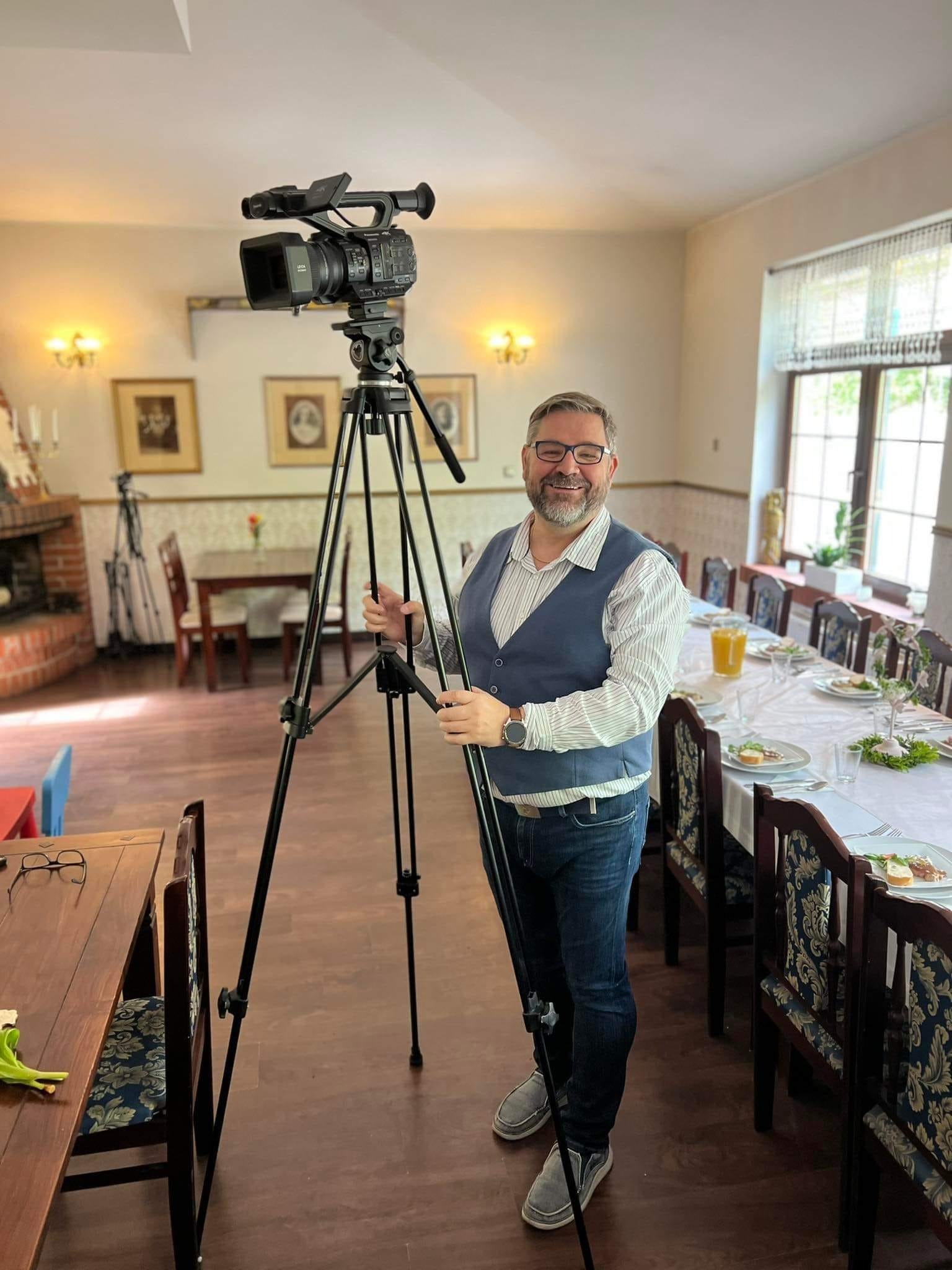 Mężczyzna w okularach i kamizelce stoi za kamerą wideo na statywie w sali z nakrytym stołem, gotowym do filmowania uroczystości.