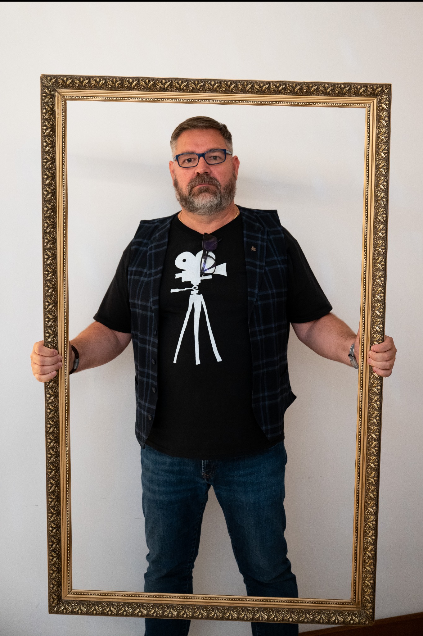 Mężczyzna z brodą i okularami, ubrany w koszulkę z rysunkiem kamery, trzymający ozdobną, złotą ramę.