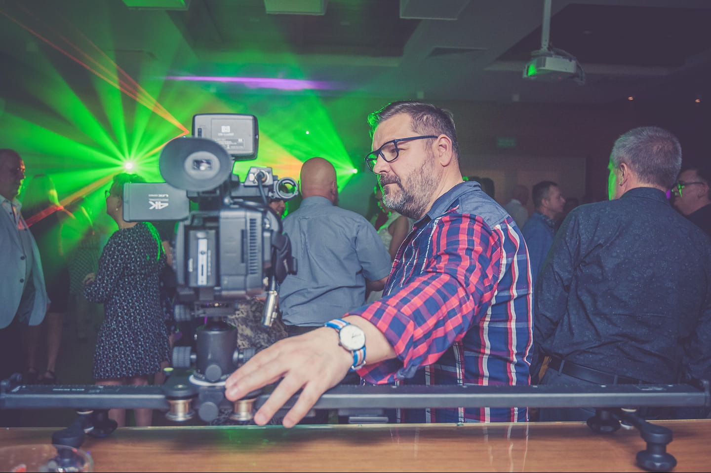 Operator kamery z brodą w okularach, w koszuli w kratę, obsługuje profesjonalny sprzęt na imprezie oświetlonej laserami.