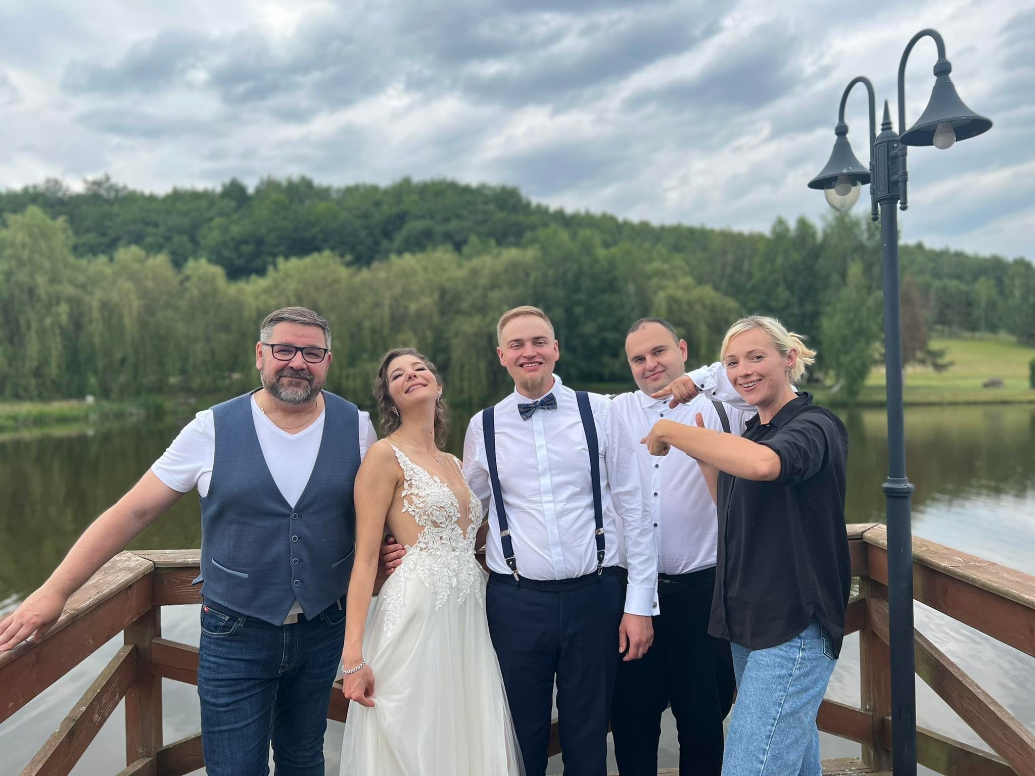 Panna młoda w koronkowej sukni i pan młody z muchą, pozujący z gośćmi na drewnianym pomoście z widokiem na jezioro i las, pochmurne niebo.