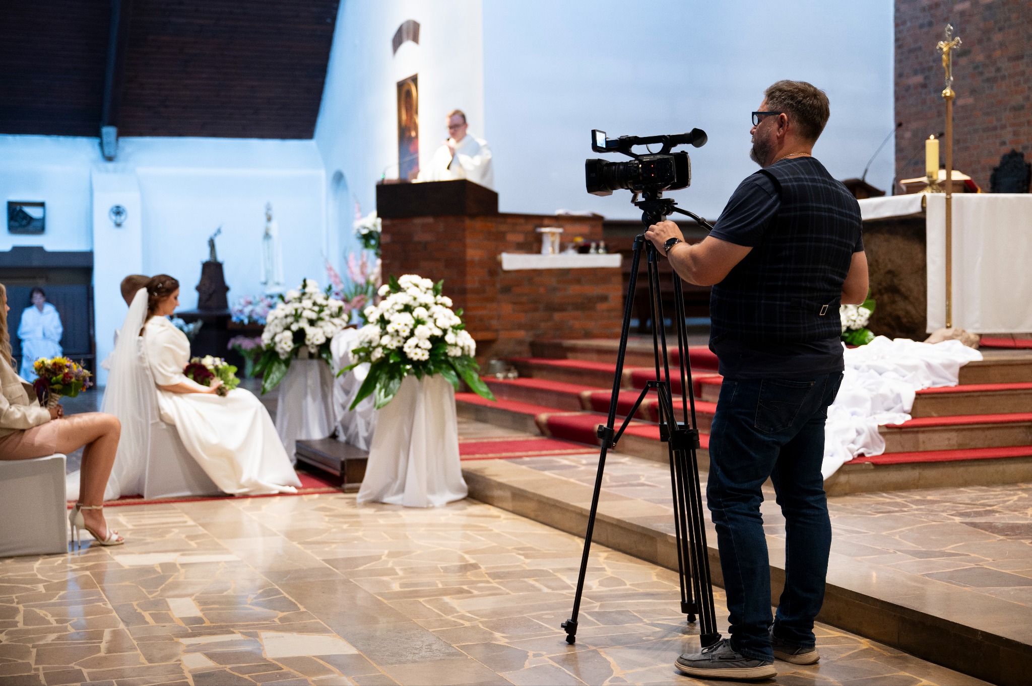 Wideofilmowanie ceremonii ślubnej w kościele: kamerzysta z kamerą na statywie filmuje parę młodą siedzącą przed ołtarzem, w tle ksiądz podczas mszy.