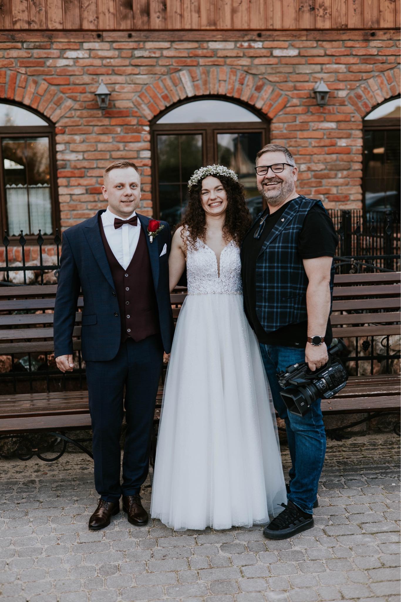Panna młoda w wianku i pan młody w bordowej kamizelce pozują z uśmiechem obok osoby trzymającej profesjonalną kamerę wideo, na tle ceglanej ściany z oknami i drewnianej ławki.