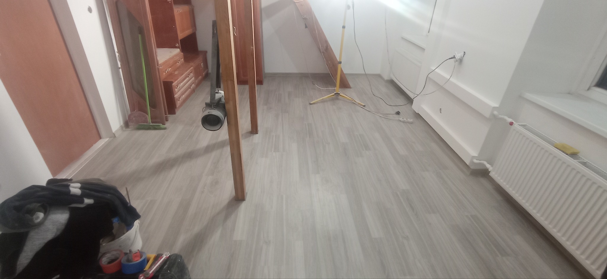 Świeżo położona podłoga z paneli winylowych w jasnym odcieniu szarości, widoczny grzejnik, lampa budowlana na trójnogu oraz narzędzia malarskie w rogu pomieszczenia.