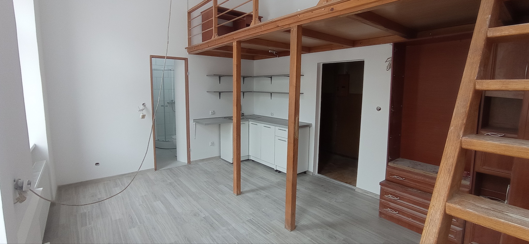 Wnętrze pokoju z antresolą wykonaną z drewna, białe ściany, szare panele podłogowe, aneks kuchenny z białymi szafkami, drabina prowadząca na antresolę i drzwi do łazienki z kabiną prysznicową.