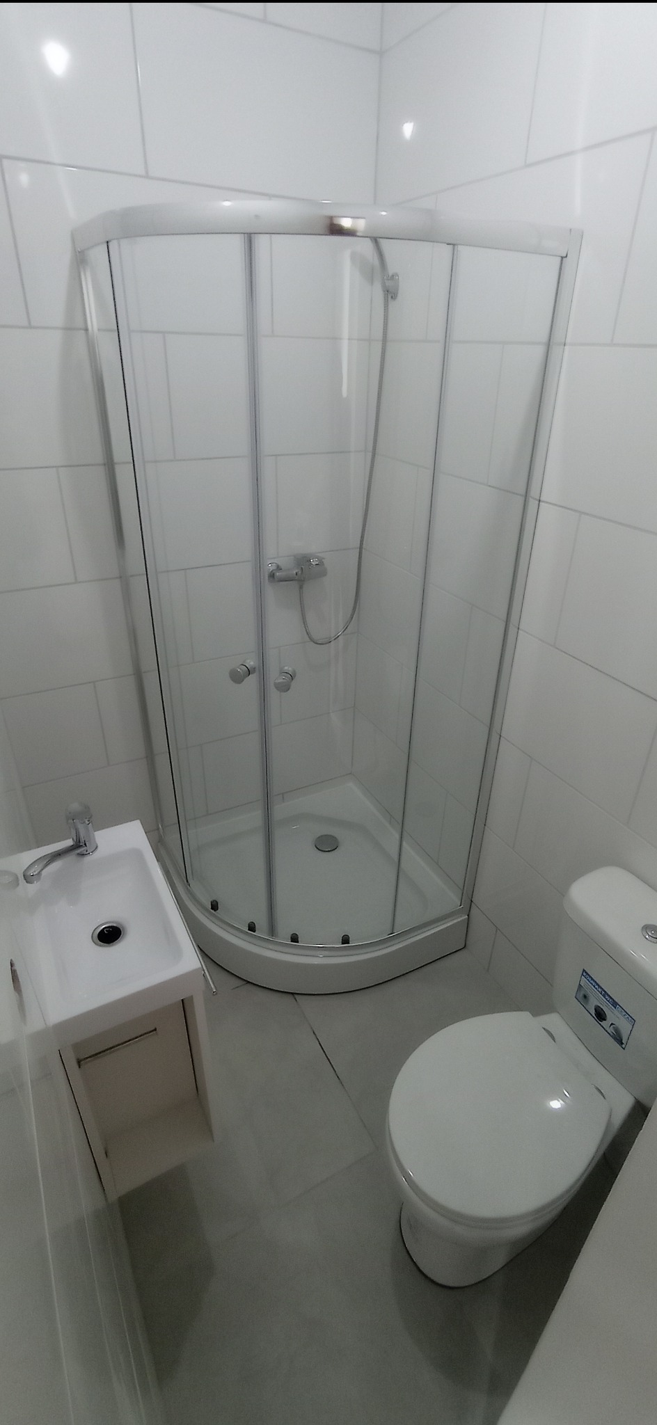 Nowoczesna łazienka z kabiną prysznicową narożną, umywalką z szafką i toaletą, wykończona białymi płytkami na ścianach i szarą podłogą.