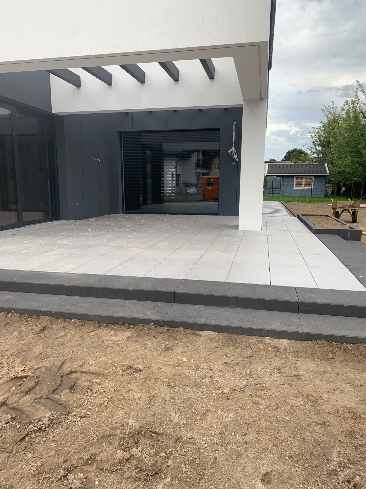 Jasnoszare płytki tarasowe z czarnymi obrzeżami na etapie układania, widoczne białe krzyżyki dystansowe, nowoczesny dom z szarymi elementami w tle, przygotowany teren pod trawnik.