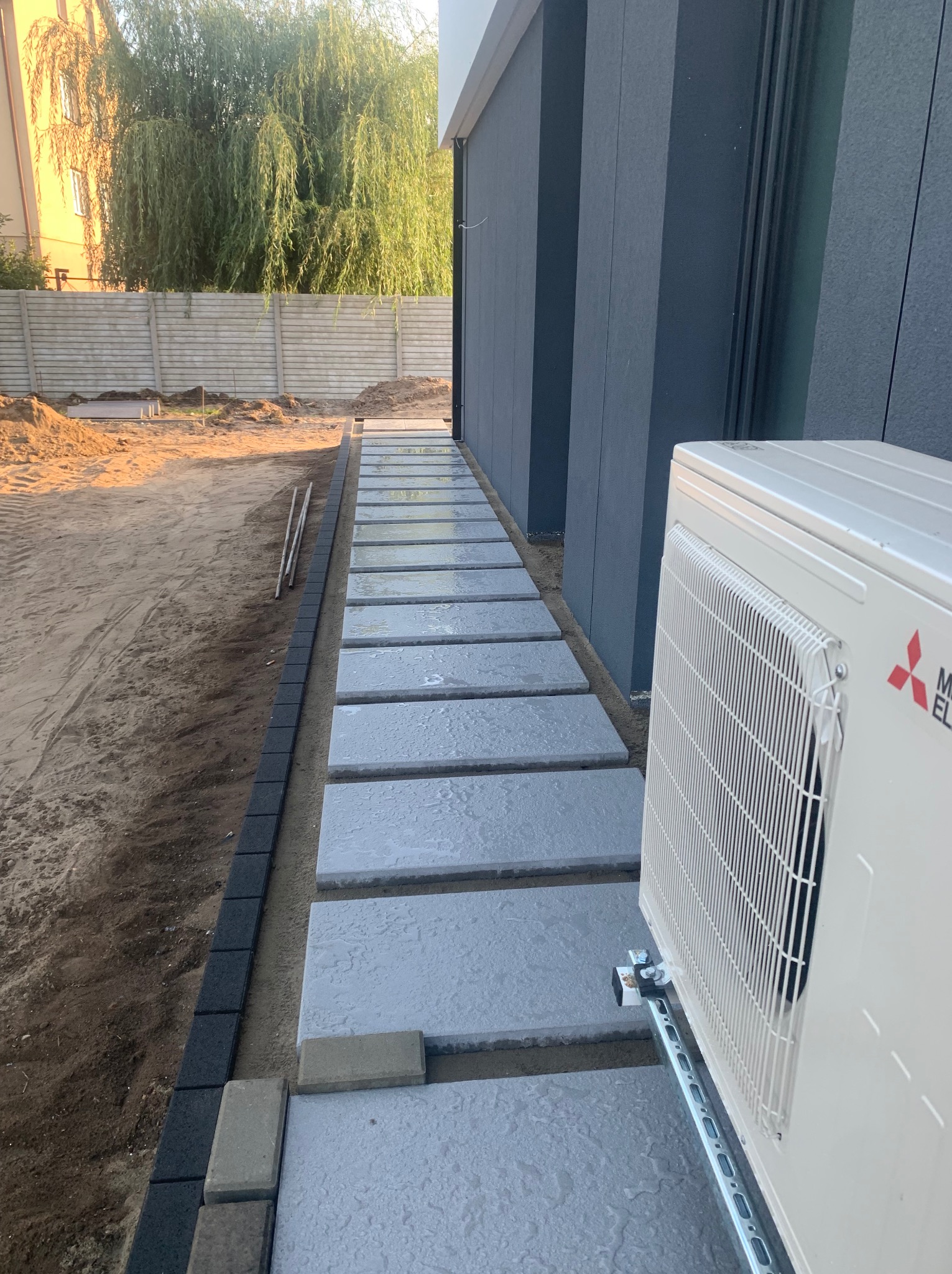 Ścieżka z prostokątnych płyt betonowych prowadząca wzdłuż ciemnej elewacji budynku, obrzeżona ciemną kostką, z widocznym klimatyzatorem na metalowej konstrukcji i przygotowanym terenem pod trawnik...