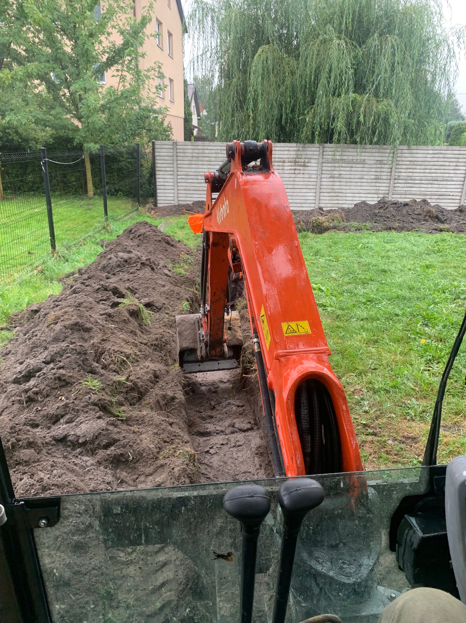 Pomarańczowa mini koparka Kubota w trakcie wykopu wąskiego rowu w ziemi na trawiastym terenie, widok z wnętrza kabiny operatora na wykopany rów, z tyłu ogrodzenie z betonowych płyt i budynek.