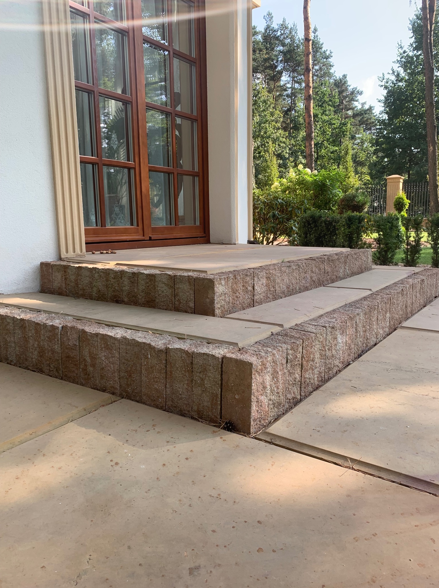 Trzy stopnie schodów wejściowych do domu wyłożone kostką granitową w odcieniach beżu i różu, otoczone płytami chodnikowymi, z widokiem na drewniane drzwi i zieleń ogrodu.